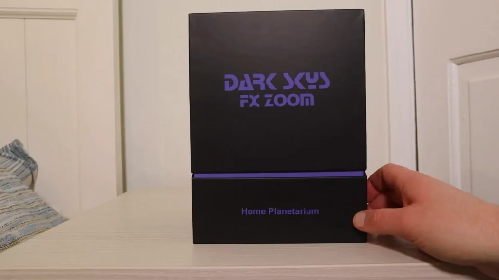 A person holds a black box labeled "DARK SKY FX ZOOM Home Planetarium" on a white table.